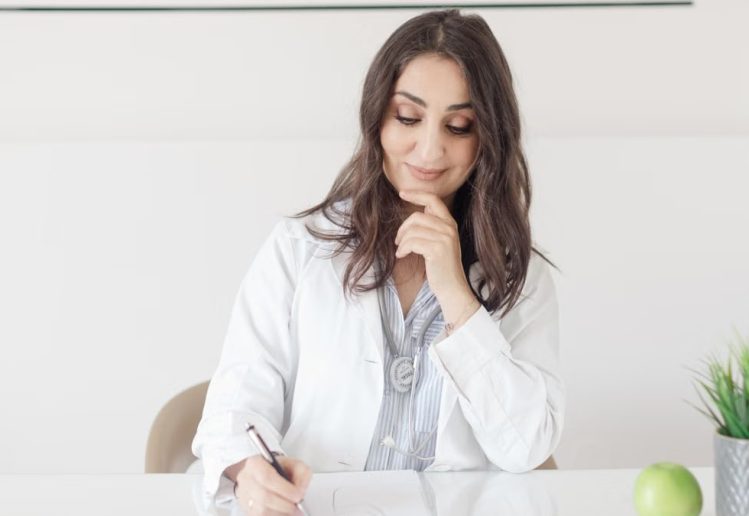 A female nutritionist in a lab coat writing notes with an apple and plant on the desk.