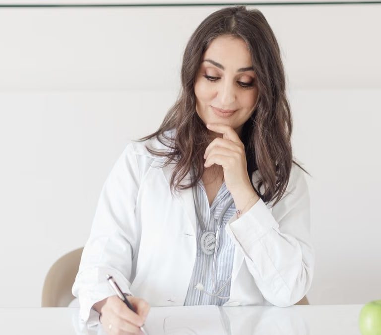 A female nutritionist in a lab coat writing notes with an apple and plant on the desk.